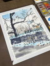 Watercolor painting showing a bare tree and stone wall dusted with snow against a pale blue sky, taped on a table next to a watercolor palette.