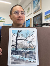 A man with glasses holds a watercolor painting of a snowy path flanked by leafless trees and a stone wall under a pale blue sky, with framed landscape paintings hanging on the wall behind him.