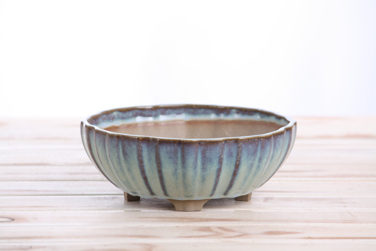 Round Willow Bonsai Ceramic Lily Pot with soft blue and brown glaze and vertical ridges, sitting on four small feet on a light wooden surface.
