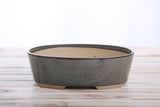 Oval ceramic bonsai pot with a muted blue-green glaze and unglazed natural beige interior, resting on a light wooden surface.