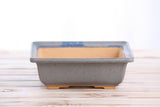 Square bonsai pot with gray exterior and glossy ocean-blue rim, featuring a tan interior and four short feet on a light wood surface.