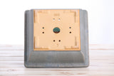 Square bonsai pot in muted gray-blue with drainage holes and a recessed beige base visible from below, placed on a light wooden surface.