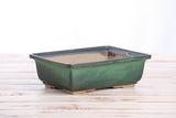 Rectangular glazed bonsai pot with an avocado green exterior and dark brown rim, resting on a light wooden surface against a white background.