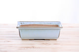 Rectangular ceramic bonsai pot in pale blue glaze with subtle brown speckles, wide flared rim, and four small feet, shown on wooden boards with white background.