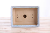 Rectangular bonsai pot with a light blue-gray rim and a natural tan interior, featuring one large central drainage hole and two pairs of smaller side holes, viewed from above on a wooden surface.