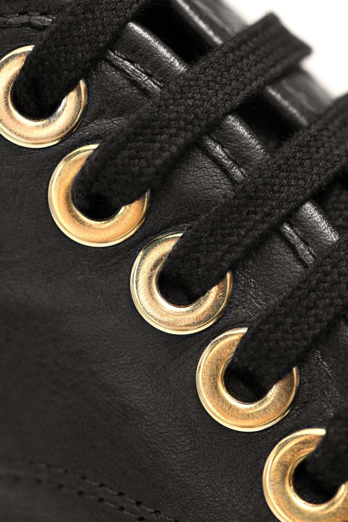 Close-up of black sneaker leather upper featuring black laces threaded through shiny gold metal eyelets with visible stitching details.