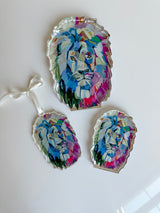 Three acrylic art pieces shaped as lion heads with colorful, abstract paint in blue, purple, pink, and green on a white background, one with a hanging ribbon.