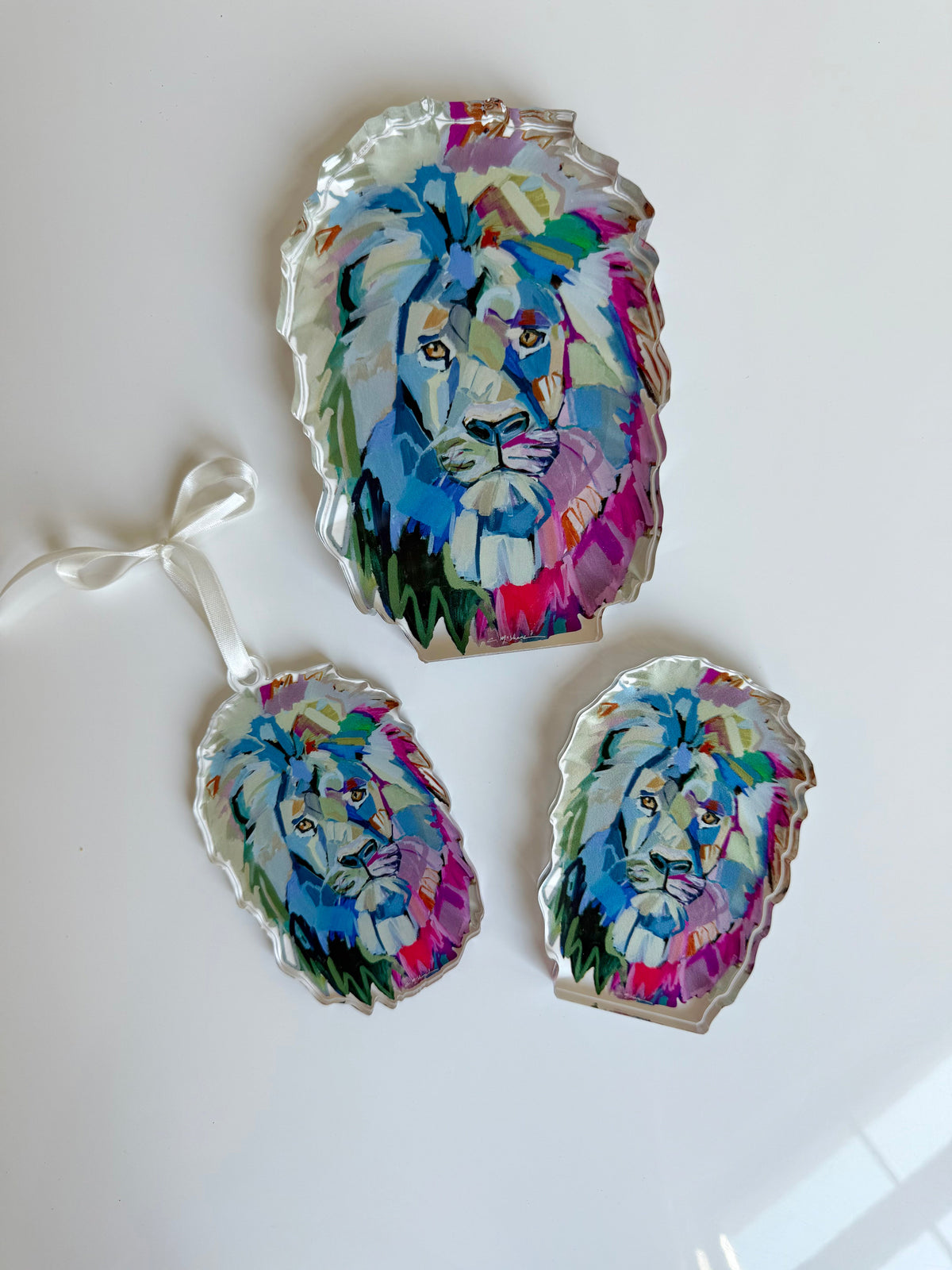Three acrylic art pieces shaped as lion heads with colorful, abstract paint in blue, purple, pink, and green on a white background, one with a hanging ribbon.