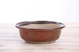 Round bonsai pot with smooth reddish-brown glaze and dark brown rim, shallow depth, sitting on a light wood surface with a white background.