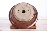 Bottom view of a round bonsai pot with a glazed reddish-brown exterior and unglazed light beige base featuring drainage holes and embossed markings, resting on a wooden stand.