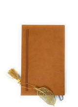 Tan suede-covered notebook with brown elastic strap, featuring a gold feather-shaped metal bookmark with matching tassel hanging from the side.