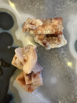 Two clusters of red apophyllite crystals with pale pink and reddish hues resting on a grey and white agate matrix with circular and banded patterns.