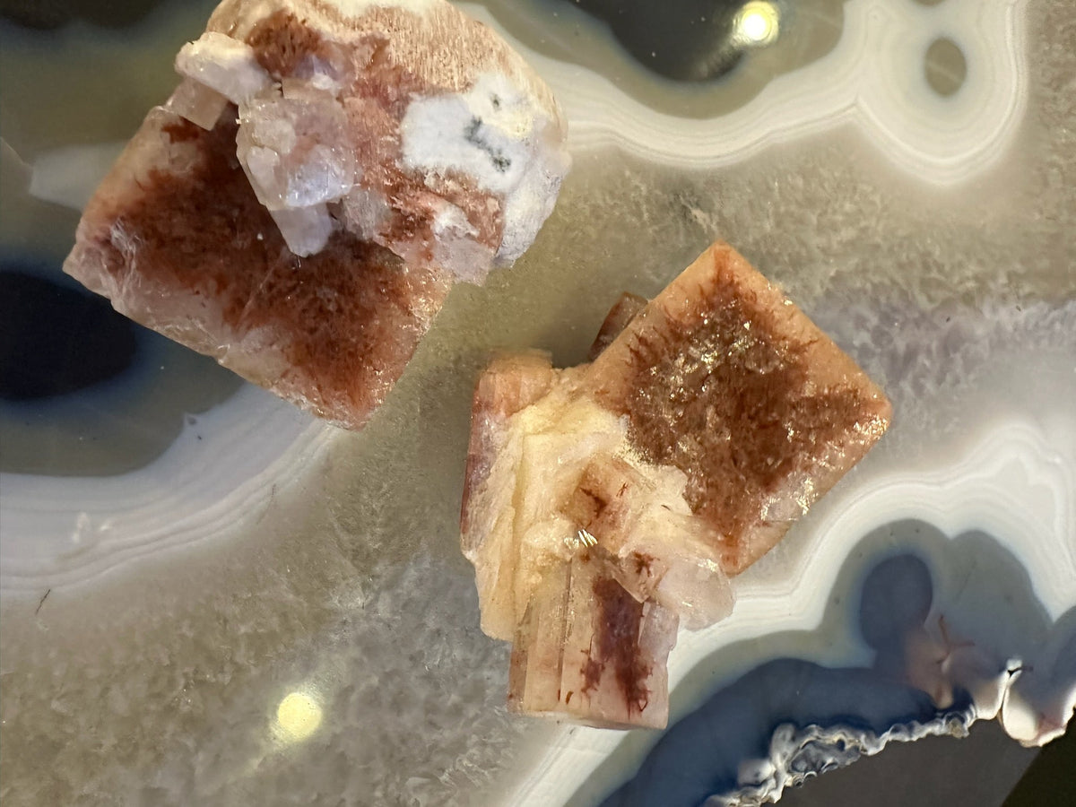 Two large red apophyllite crystals with white and translucent formations on a smooth, banded gray and blue agate surface, illuminated to highlight their textures.