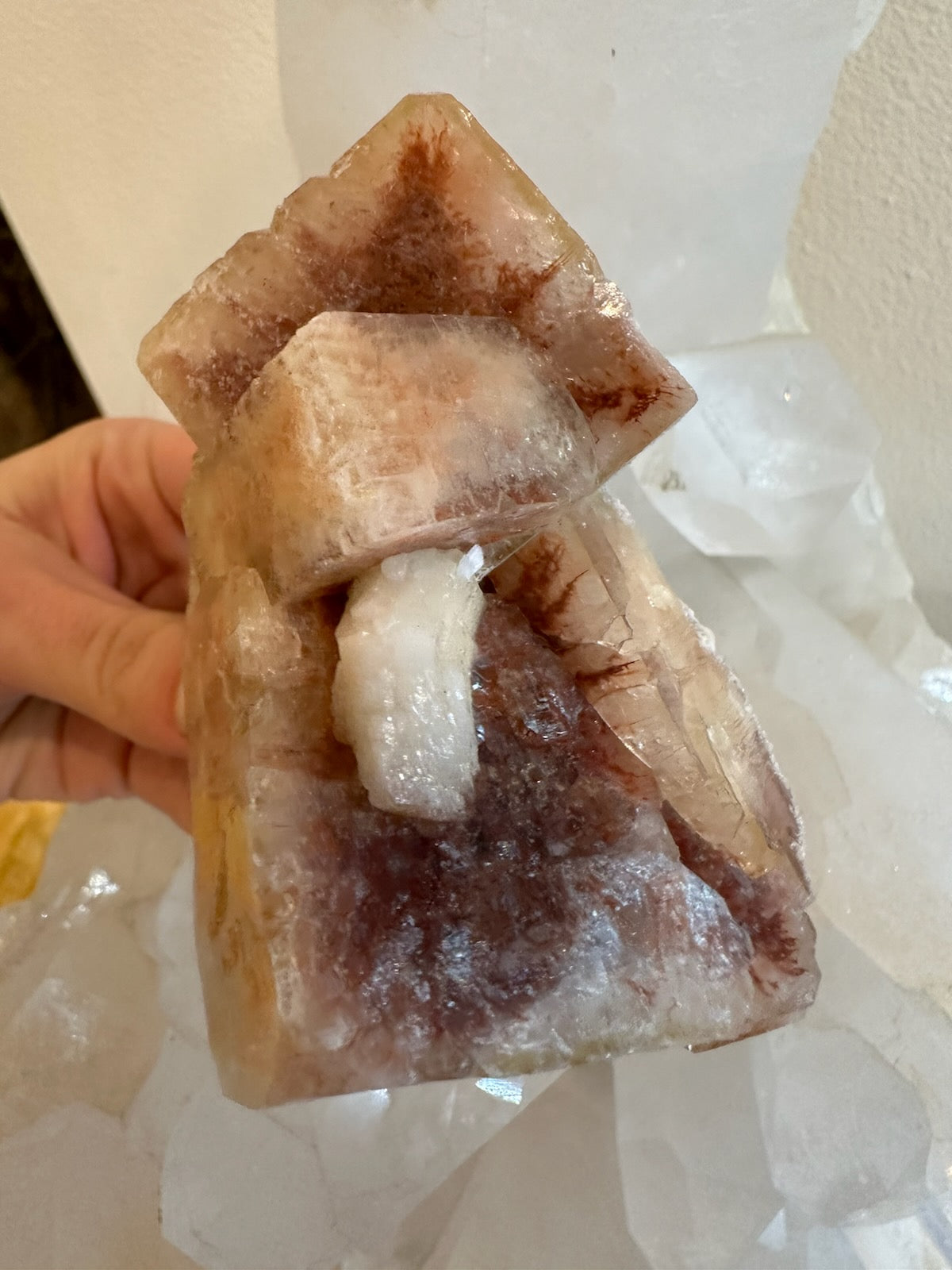 Translucent red and white apophyllite crystal cluster with cube and pyramid shapes held by a hand against a background of similar clear crystals.