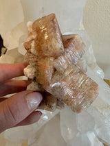 Hand holding a cluster of reddish-brown translucent apophyllite crystals with a cubic structure, set against a backdrop of larger clear crystals.