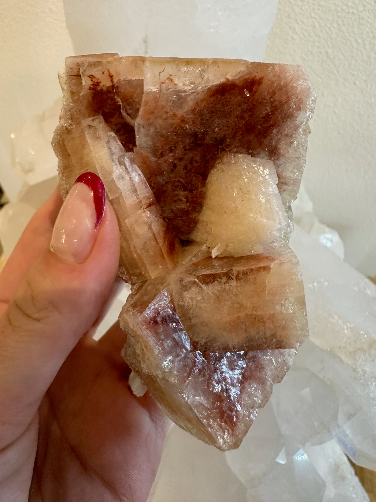 Hand holding a red apophyllite cube cluster with translucent, reddish-brown and cream crystals showing layered cubic shapes and reflective surfaces.