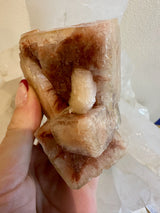 Hand holding a cluster of red and translucent apophyllite cubic crystals with varying sizes and a smooth, reflective surface under natural light.