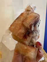 Hand holding a translucent red apophyllite cube with orange and brown tones and white stilbite crystals on its surface against a blurred neutral background.