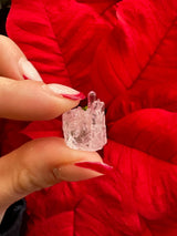 Close-up of a small, clear micro rose quartz crystal held between fingers with red-painted nails, set against a vibrant red leaf background.