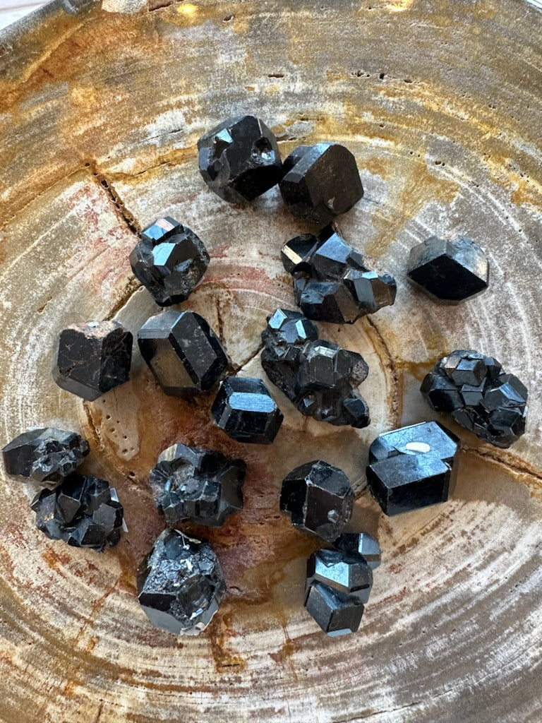 Cluster of glossy black melanite garnet crystals with varied geometric shapes and smooth to faceted surfaces, resting on a textured, circular wooden surface.