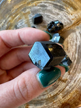Hand holding a lustrous black melanite garnet crystal with geometric facets over a wooden surface scattered with similar black crystals.
