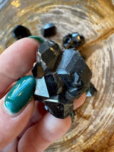 Hand holding a black melanite garnet crystal with angular, reflective facets and a rough, geometric shape against a textured wooden background with additional crystals scattered.