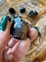Close-up of a person's hand holding a dark, angular melanite garnet crystal above a wooden surface with several similar dark crystals scattered around.