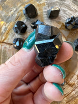 Close-up of a shiny black melanite garnet crystal held between green-painted fingernails, with several similar crystals on a textured wooden surface in the background.