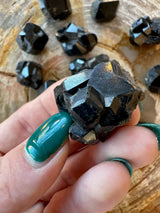 Close-up of a hand holding a shiny black melanite garnet crystal with geometric facets, surrounded by similar crystals on a wooden surface.