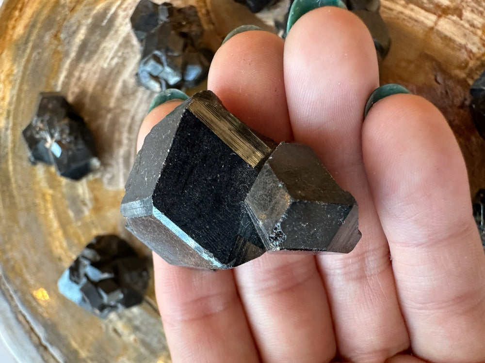 Hand holding a dark, faceted melanite garnet crystal with a geometric shape and slightly reflective surface, surrounded by similar crystals on a wooden surface.