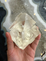 Hand holding a translucent golden calcite crystal with smooth, glassy surfaces and angular shapes against a patterned agate stone background.