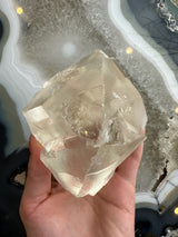Hand holding a translucent golden calcite crystal with smooth, angular surfaces and internal fractures, against a gray and white banded agate backdrop.