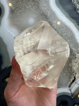 Hand holding a translucent golden calcite crystal with angular facets, displayed against a patterned gray and white agate slab background.