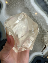 Hand holding a translucent golden calcite crystal with natural facets, against a background of banded gray and white agate stone slices.