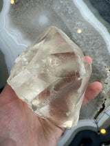 Hand holding a large, clear golden calcite crystal with smooth, angular faces and internal fractures, against a gray and white banded geode background.