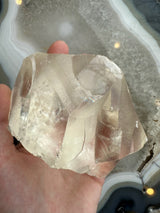Hand holding a large, translucent golden calcite crystal with angular facets and internal striations, over a patterned agate slice background.