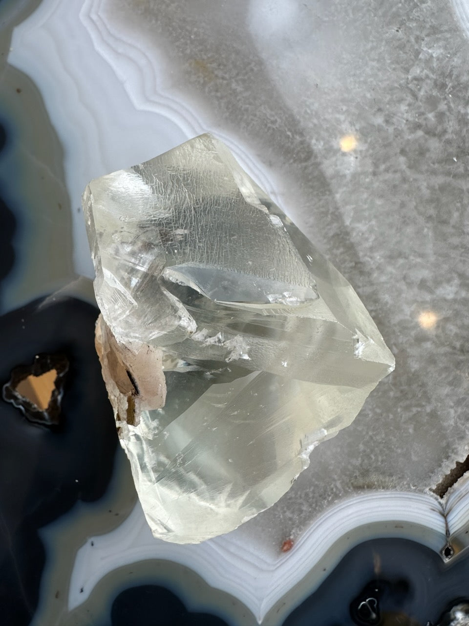 Translucent pale golden calcite crystal with angular facets resting on a polished agate slab featuring concentric white and black bands and a dimpled texture.