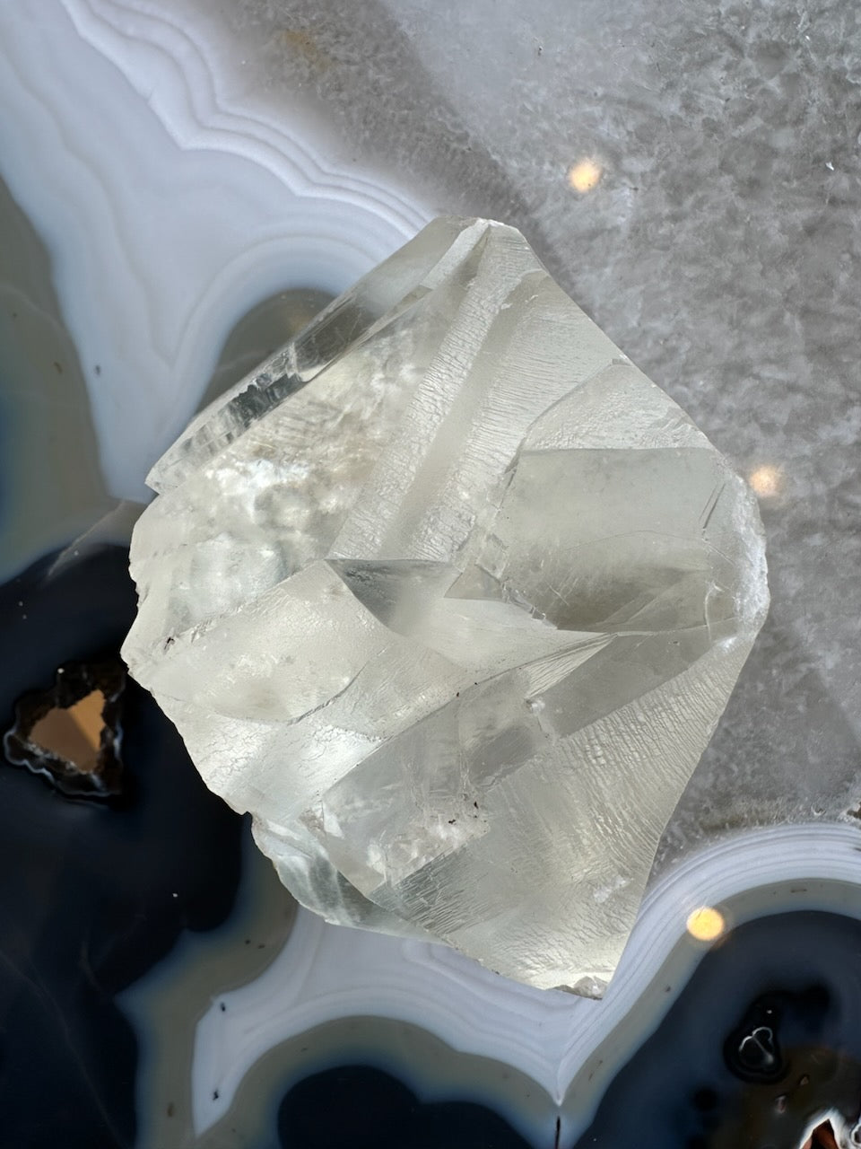Translucent pale yellow calcite crystal with geometric facets, resting on a patterned agate surface featuring white and dark concentric bands.