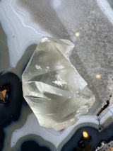 Translucent golden calcite crystal with polished facets resting on a banded agate surface featuring grey, white, and black concentric patterns with sparkly texture.