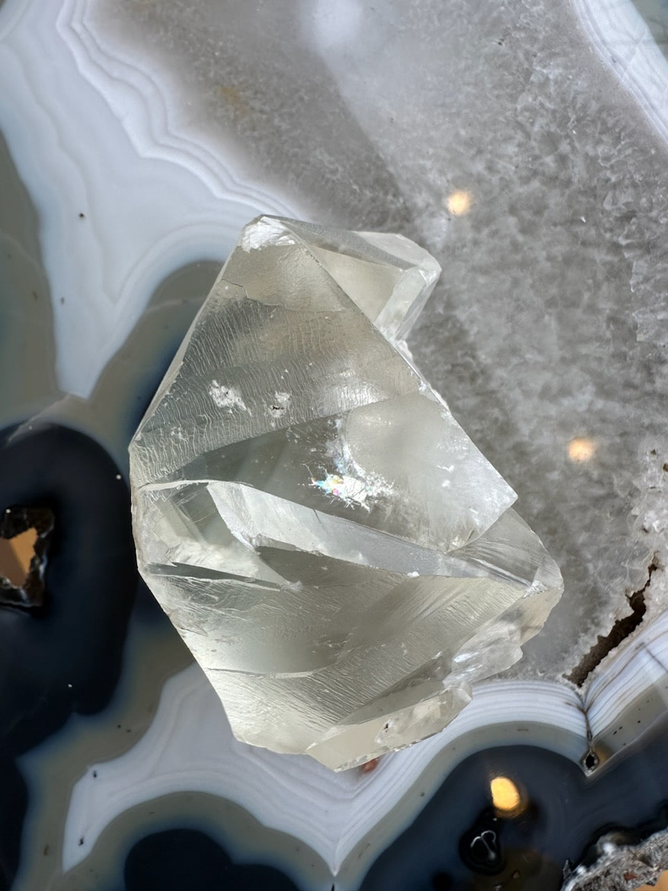 Translucent golden calcite crystal with polished facets resting on a banded agate surface featuring grey, white, and black concentric patterns with sparkly texture.