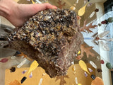 Hand holding a large, irregularly shaped brown rock densely covered in dark reddish-brown garnet crystals, with a decorative background of dried leaves and crystals.