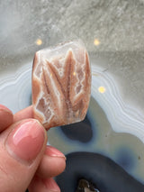 Polished Apple Valley agate pseudomorph cabochon with peach and cream banding, held between fingers against a background of a blue and white geode slice.