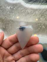 Smooth, polished, translucent moss agate cabochon with a pointed shape held between fingers over a patterned stone surface with white and gray tones.