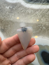 Fingers holding a translucent, cone-shaped moss agate cabochon with subtle black inclusions, shown over a glossy agate slab with concentric patterns.
