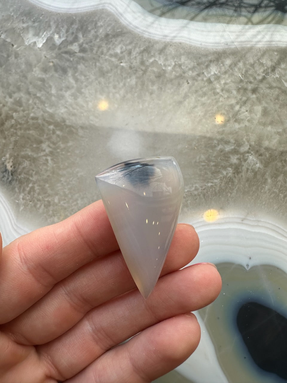 Hand holding a polished, translucent moss agate cabochon shaped like an elongated cone with faint internal inclusions against a textured gray and white stone background.