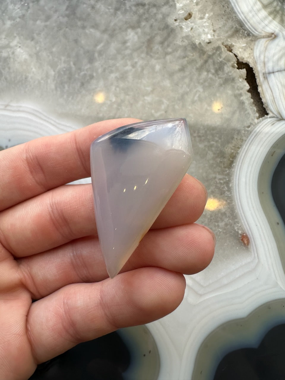 Hand holding a polished translucent Moss Agate cabochon with smooth, curved surfaces over a gray and white banded agate background.
