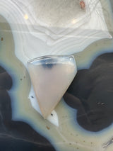 Translucent triangular moss agate cabochon with smooth polished surface resting on a banded agate slab featuring concentric rings of grey, white, and brown hues.