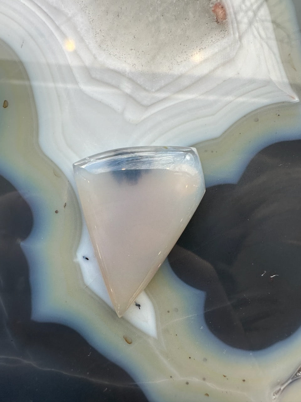 Translucent triangular moss agate cabochon with smooth polished surface resting on a banded agate slab featuring concentric rings of grey, white, and brown hues.