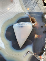 Polished translucent moss agate cabochon with soft white and light blue hues resting on a polished geode surface featuring concentric bands and earthy brown inclusions.