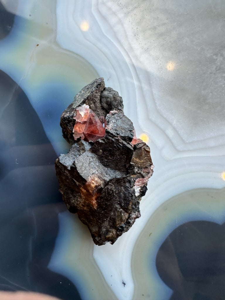 Dark matrix rock featuring translucent pink rhodochrosite crystals with geometric shapes, resting on a smooth, banded agate surface with white and blue-gray layers.